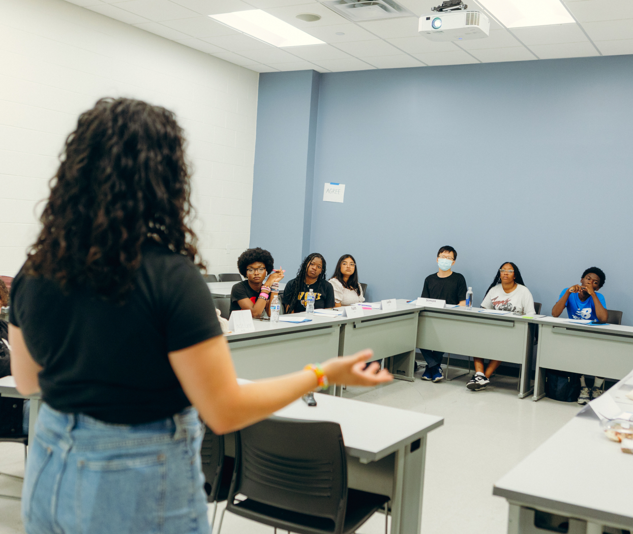 A teacher is speaking in front of a group of students in a classroom with hand gestures. Students are sitting around U-shaped desks, listening to the teacher.