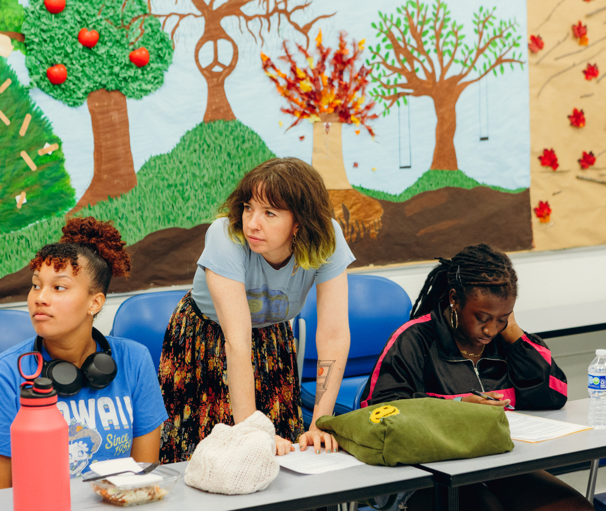 Two students sit at a table while a teacher stands between them. The teacher and one of the students looks to the left of the frame.